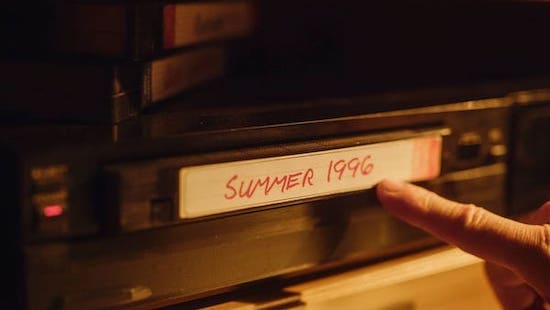 A hand points to a VHS tape labeled "Summer 1996" on a shelf, with other tapes stacked nearby.