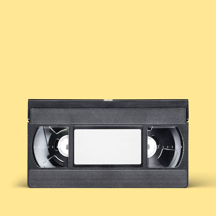 A black VHS tape with a blank label is set against a solid yellow background.